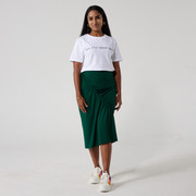 A south asian model is smiling at the camera. She is wearing a forest green ruched skirt with a white t shirt and white sneakers. Her hands are relaxed to her sides. 