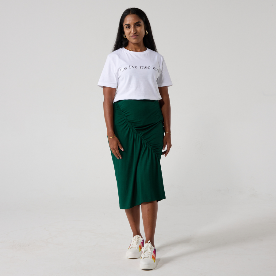 A south asian model is smiling at the camera. She is wearing a forest green ruched skirt with a white t shirt and white sneakers. Her hands are relaxed to her sides. 