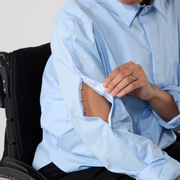 CLose up of a pale blue long sleeve shirt that has openings in the sleeves that use popper tape to open and close. A black wheelchair is just visible on the left of the image.