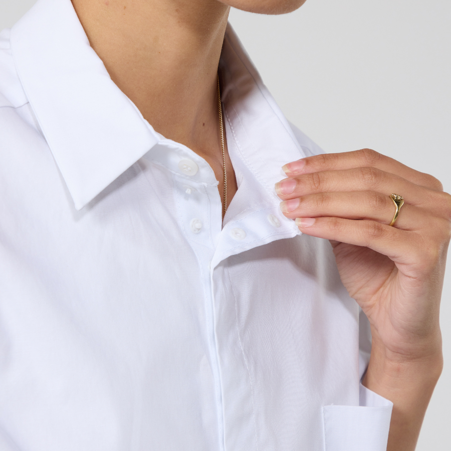 Close-up on the collar, button closure and popper tape on the white short sleeve hi-lo shirt. It is modelled on a non-binary model with mixed heritage. She is holding one of the collar flaps with one hand. 