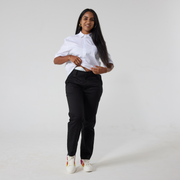 A South asian woman with long back hair is wearing black twill trousers in regular fit paired with a white shirt and sneakers with rainbow patches. She is lifting up her shirt hem to show her stoma bag and the accessibility of the trousers. 