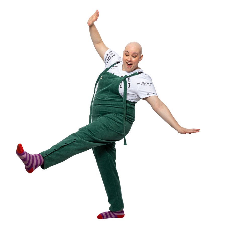 Laura, a white woman with alopecia, is standing one leg, smiling and looking down at the floor. She is wearing green dungarees with a white t shirt underneath reading "disabled is not a bad word" with stripy purple and pink socks