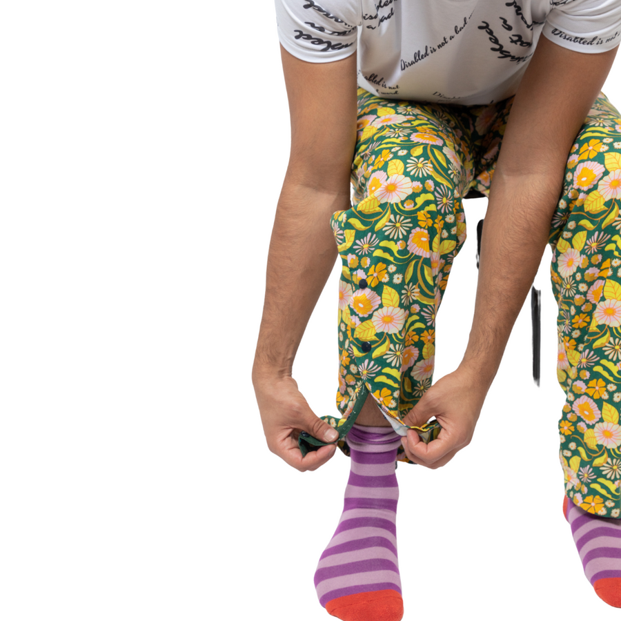 A close up of the popper features on the bottom of the green and yellow bright print trousers. The model is sitting on a black stool and it also wearing purple and pink stripy socks