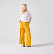 A white woman with blonde hair poses standing, looking away from the camera. She is wearing a white Jersey wrap top paired with yellow culottes and her own boots. She is standing with her hands in her pockets against a light grey background.