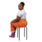 Glynis, a black woman with an arm limb difference, is wearing pink and yellow vibrant , high waisted trousers with a white tshirt with the text slogans "Disabled is not a bad word" .She is sitting on a black stool with her back to camera and looking over her shoulder. Her trousers are turned up at the bottom with purple and pink stripy socks