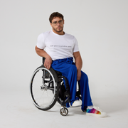 A male model of mixed heritage is looking at the camera. He is wearing Royal blue jersey culottes in regular fit paired with a white t-shirt and white sneakers. He is sat in a manual wheelchair with one of his hands resting against the side of the wheelchair and the other in his lap.