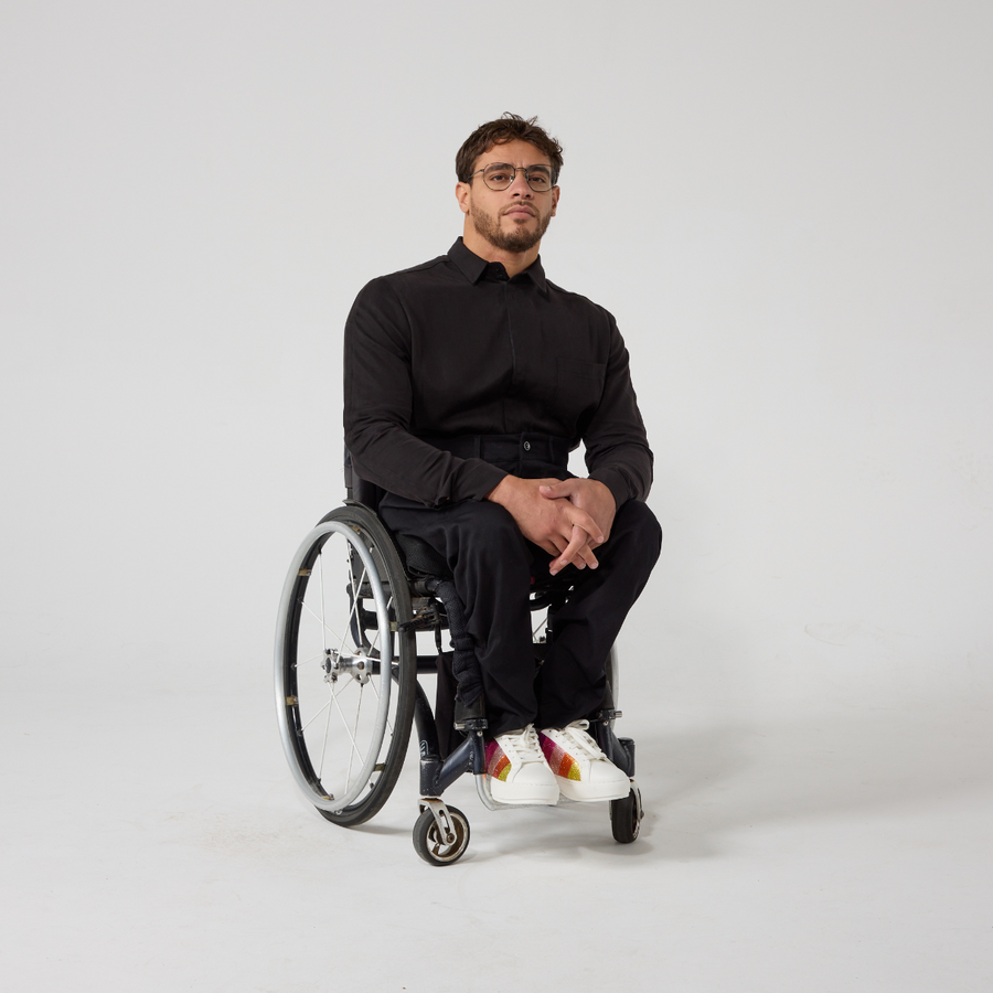 Moatez is a male presenting mixed heritage model wearing a black shirt and black trousers with white sneakers that have sparkly rainbow accents on the side. He is a manual wheelchair user and has black wire frame glasses on. He's in front of an off white back ground.
