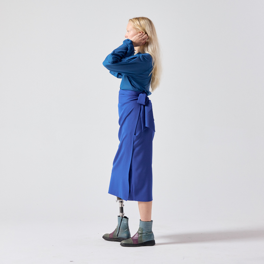 Side view of a white woman with long blonde hair. She is wearing a cobalt blue wrap skirt with a teal shirt and her own boots. She is standing with her hands tucking her hair behind her ears.