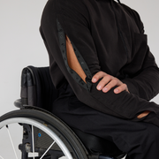 Close up shot of a black shirt that has sleeve openings via popper tape. Model is a mixed heritage wheelchair user.