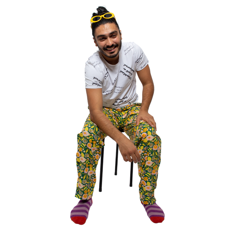 Moeed is a south Asian man, with black hair in bun and yellow sunglasses on his head. He is sitting on a black stool against a white background wearing seated floral yellow and green printed trousers with a white t-shirt with the slogan "Disabled is not a bad word" written across it. He is smiling at the camera.