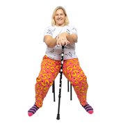 Sylvia, a white woman, is wearing pink and yellow vibrant , high waisted trousers with a white tshirt with the text slogans "Disabled is not a bad word" .She is sitting on a black stool and is also wearing purple and pink stripy socks. She is smiling at the camera and holding her arms out in front of her holding a black mobility aide with white stars on it