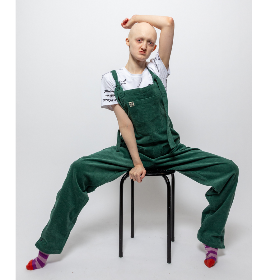 Hannah, a white woman, is sitting on a black stool with her legs apart and her arm resting on her head. She is wearing green dungarees with a white t shirt underneath reading "disabled is not a bad word" with stripy purple and pink socks