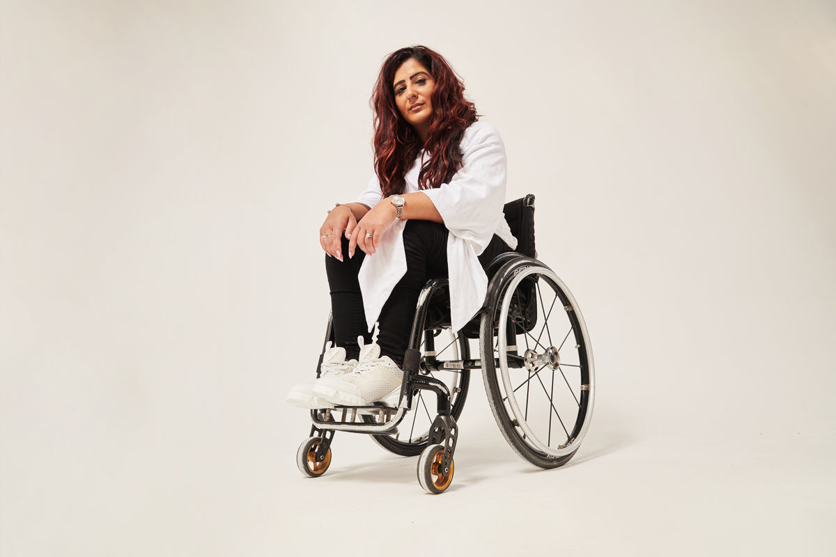 A woman with long brown hair is looking into the camera. Her arms are resting on her knees. She is wearing a white adaptive wrap top with winged sleeves. She is wearing black seated adaptive trousers. She is sitting in a manual wheelchair. 