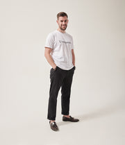 A smiling white man with dark brown hair and dark stubble looks at the camera.  His hands are in his pockets. He is wearing a white t-shirt with Unhidden on it and black trousers. He has brown brogues on.