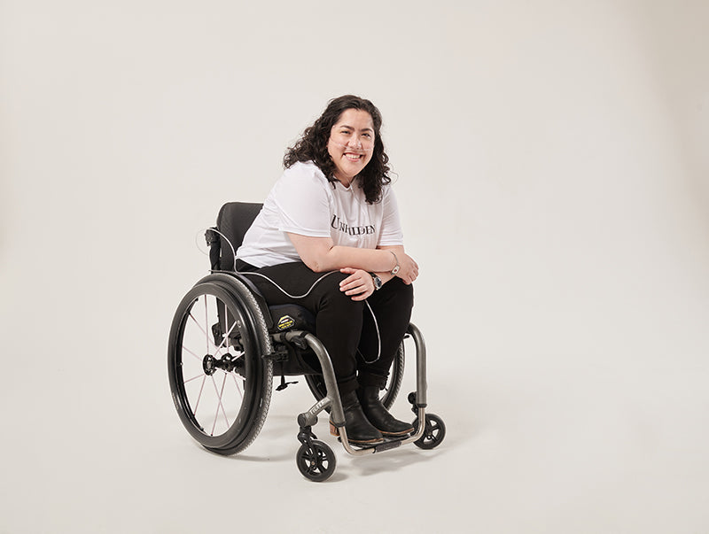 A smiling woman with curly dark brown hair looks at the camera. She is wearing a white t-shirt with unhidden written on it, and black trousers. Her arms are crossed in her lap and she leans slightly forward. She is sitting in a manual wheelchair and wearing an oxygen tube.