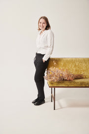 A white woman smiles at the camera. Her hands are in her pockets. She is wearing a white shirt with concealed openings at her arms and chest. A tube rests on her stomach. The back ground is white, she is leaning on a. crushed velvet, moss coloured sofa. There is a bunch of dried flowers on the sofa next to her.
