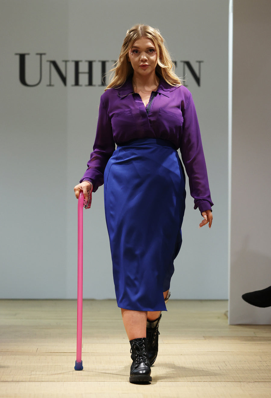 Female presenting model on a runway. They are a white woman with wavy long blonde hair. Wearing a purple chiffon shirt with long sleeves under a blue wrap skirt. They are using a hot pink walking stick and have chunky black boots on. Behind them is a white back drop with Unhidden in black text visible.