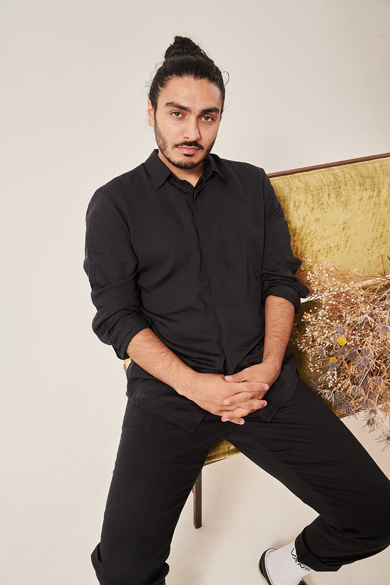 A south Asian man with long dark hair in a bun looks serenely in to the camera. He is wearing a bamboo silk shirt rolled to the elbows, and black trousers that are rolled at the hem. He is sitting on a crushed velvet moss coloured sofa, with some dried flowers on the seat to his right. The background is white.