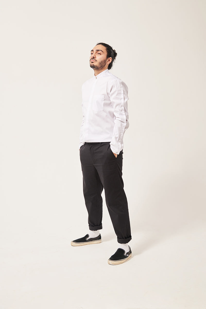 A south Asian man stares moodily off camera. His hair is tied up, his hands are in his pockets. He is wearing a white shirt with a seam down the sleeve and a chest pocket. The collar is a mandarin style with a shell button, and the black trousers are rolled at the hem showing his white socks and plimsoll shoes. The background is tones of grey.