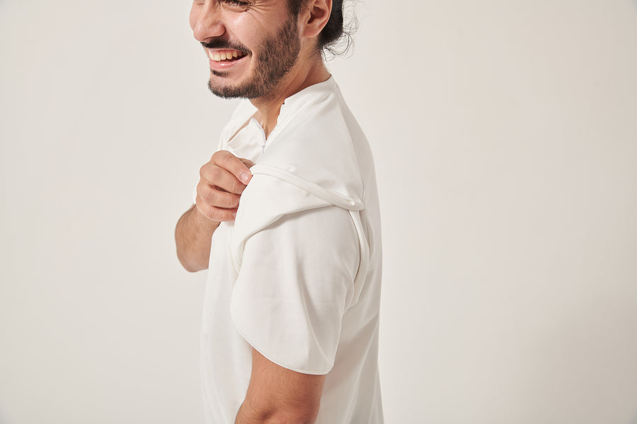 A south Asian man is laughing off camera, the top of his head is cropped. He is pulling up part of the wrap sleeve on his white t-shirt. The background is white. There is a zip pulled open on his chest for port access.