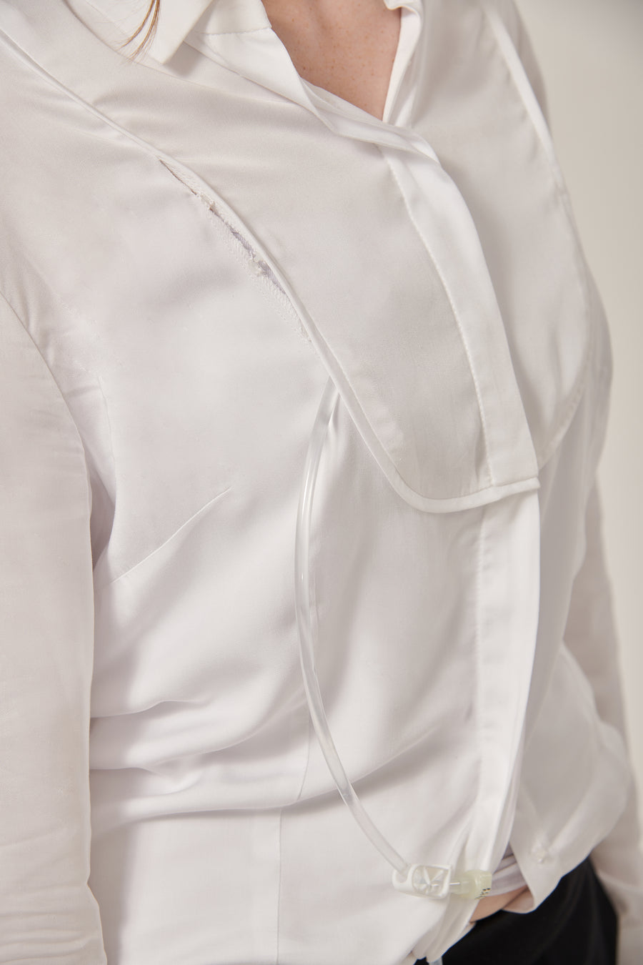 Close up of a woman's white shirt. There is a bib styling detail that conceals a chest opening. A tube comes from the woman's waist and loops back in at the chest.