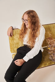 A woman with dark blonde curly hair is laughing and look off to the left of the frame. She is wearing black glasses and is reclining on a crushed velvet moss coloured sofa. The tip of a bunch of dried flowers is on the seat beside her. She is wearing a white shirt with a concealed sleeve opening and black trousers adapted for wheelchair users. There is a wrap the front.