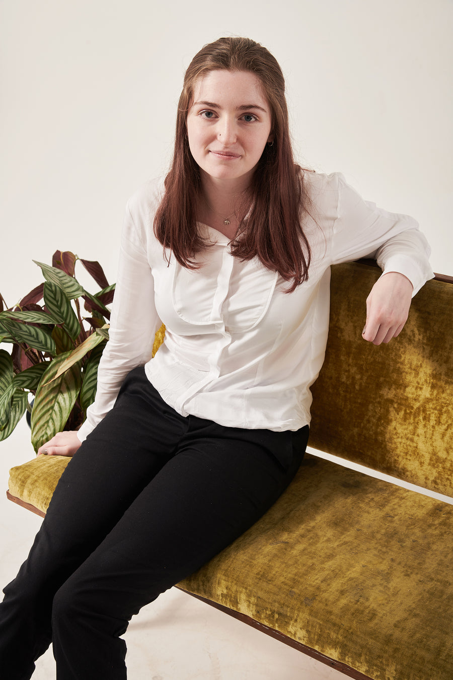 A woman with dark brown hair in a half up half style smiles in to the camera. She is wearing a white shirt with a bib seam detail and black trousers. She is siting on a crushed velvet sofa that is moss coloured. There is a plant on the floor just behind her. The background is white.